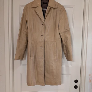 Beige kappa från Rock'n Blue - Snygg beige kappa från Rock'n Blue i klassisk design. Kappan har långa ärmar och knappar framtill. Perfekt för en stilren look under höst och vår.