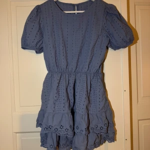 Blå klänning med volangdetaljer - Charmig blå klänning med korta ärmar och volangdetaljer. Klänningen har ett broderat mönster och en elastisk midja för en bekväm passform. Perfekt för sommarens varma dagar.