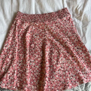 Rosa blommig kjol - Söt rosa kjol med blommigt mönster i vitt och svart. Kjolen har en elastisk midja för bekväm passform och är perfekt för en somrig look. Materialet är lätt och luftigt, vilket gör den idealisk för varma dagar.