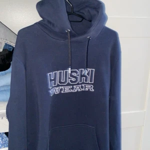 Mörkblå hoodie från Huski Wear - Säljer en mörkblå hoodie från Huski Wear med tryck på bröstet. Den har en justerbar huva med snören och en stor ficka framtill. Perfekt för kyliga dagar!