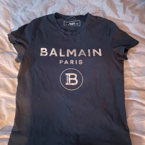 Svart t-shirt från Balmain - Snygg svart t-shirt från Balmain med guldtryck av märkesnamnet och logotypen på framsidan. Perfekt för en stilren look. Äkta 
