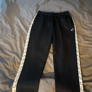 Svarta mjukisbyxor från Fred Perry  - Snygga svarta mjukisbyxor med elastisk midja och logotypband längs sidorna. Perfekta för en avslappnad stil. Byxorna har en normal passform och är tillverkade i ett mjukt material för extra komfort.