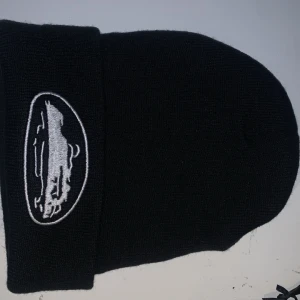 Svart stickad mössa med broderad logga - Snygg svart stickad mössa med en vit broderad logga framtill. Perfekt för att hålla värmen under kyliga dagar. Enkel och stilren design som passar till det mesta.