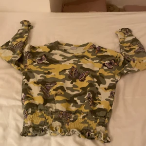 Camouflage tröja med fjärilar från Lindex - Säljer en cool tröja från Lindex med ett unikt camouflagemönster i grönt och gult, dekorerad med fjärilar. Tröjan har långa ärmar och en croppad design med volangdetaljer vid nederkanten. Perfekt för den som vill sticka ut!