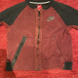 Bordeaux och svart kofta/tröja från Nike - Snygg bordeaux och svart kofta/tröja från Nike med dragkedja och logga på bröstet. Koftan har långa ärmar och ribbade muddar. Perfekt för en stilren look.