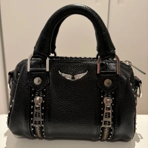 Zadig&Voltaire väska med nitar - Snygg svart handväska från Zadig i skinn med coola detaljer som nitar och dragkedjor. Väskan kan både bäras crossbody eller i handen.