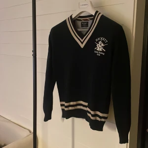 Hackett London v-ringad tröja - Säljer en stilren mörkblå v-ringad tröja från Hackett med broderad logga på bröstet. Tröjan är i storlek S. Tillverkad i bomull och perfekt för en klassisk look.