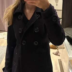 Svart kappa med knappar - Snygg svart kappa med dubbelknäppning och långa ärmar. Perfekt för kyligare dagar och ger en stilren look. Passar bra till både vardag och mer uppklädda tillfällen.