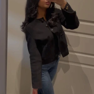 Svart kappa - Snygg svart kappa med knappar framtill. Den har en klassisk design med långärmade ärmar och en normal passform. Perfekt för en stilren look.