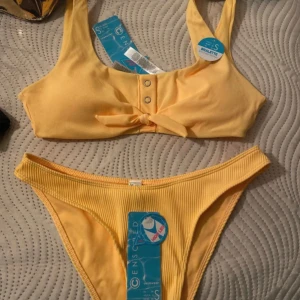 Gul ribbad bikini med knyt - Trendig gul bikini från Censored i ribbat material. Överdelen har knyt och dekorativa knappar framtill, medan underdelen är i en klassisk tanga-stil. Perfekt för soliga dagar på stranden!