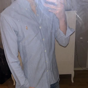 Blå skjorta från Ralph Lauren - Snygg blå skjorta från Ralph Lauren med klassisk krage och broderad logga på bröstet. Skjortan har långa ärmar och knappar framtill. Perfekt för en stilren look.