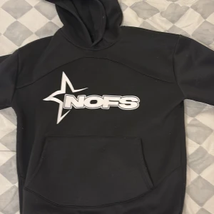 Svart hoodie med NOFS-logga - Snygg svart hoodie med stor vit NOFS-logga på framsidan. Tröjan har en klassisk känguruficka och en bekväm huva. Perfekt för en avslappnad stil.