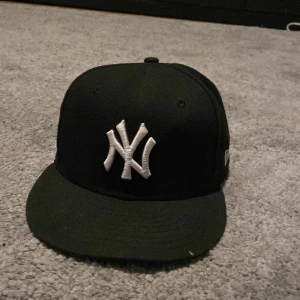 Svart keps från New Era - Snygg svart keps från New Era med ett broderat vitt NY-logotyp framtill. Kepsen har en klassisk design och är perfekt för vardagsbruk. Den är tillverkad i ett slitstarkt material och har en bekväm passform.