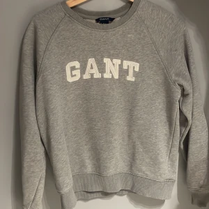 Grå sweatshirt från GANT - Säljer en stilren grå sweatshirt från GANT med deras logga i vitt på bröstet. Tröjan har långa ärmar och ribbade muddar vid ärmslut och nederkant. Perfekt för en avslappnad stil.Har några små fläckar men inget man tänker på.