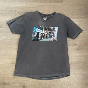 Supreme tshirt  - Säljer en grå t-shirt från Supreme med ett coolt tryck av en stadsmiljö på framsidan. T-shirten har en klassisk rund hals och korta ärmar. Perfekt för en avslappnad stil.