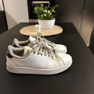 Vita sneakers från Adidas - Säljer ett par klassiska vita Adidas sneakers med perforerade detaljer på sidorna och mörkblå hälflik med Adidas-logga. Skorna har snörning och en stilren design som passar till det mesta. Använda ca 2 månader