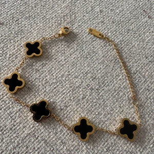 Armband - Elegant guldarmband med fem svarta klöverformade hängen. Armbandet har en justerbar kedja och ett smidigt lås. Perfekt för att ge en stilren touch till din outfit.