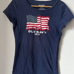 Mörkblå t-shirt från Old Navy - Säljer en mörkblå t-shirt från Old Navy med ett tryck av den amerikanska flaggan och texten '2012'. Perfekt för en avslappnad stil. T-shirten är kortärmad och gjord i bomull.