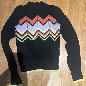 Svart tröja med zigzag-mönster från River Island S - Snygg svart tröja från River Island med färgglatt zigzag-mönster i rött, lila och gult. Tröjan har långa ärmar och en hög krage, perfekt för att hålla värmen. Den har ribbade muddar vid ärmslut och nederkant för en bekväm passform.