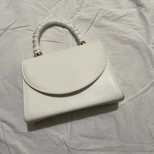 Vit handväska med kedja - Snygg vit handväska med en elegant kedja som axelrem. Väskan har ett stilrent och minimalistiskt utseende med ett handtag som är täckt av ett rynkat material. Perfekt för att ge en touch av elegans till din outfit.