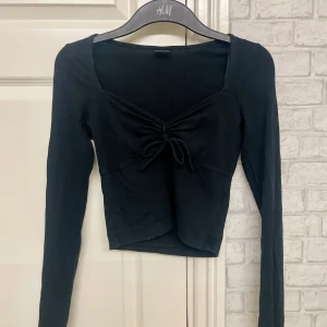 Svart långärmad topp från Gina Tricot - Snygg svart långärmad topp från Gina Tricot med knytning framtill och slits i slutet av ärmarna. Toppen har en croppad design och är perfekt för en stilren look. Passar bra till både jeans och kjol.🌺
