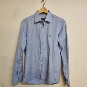 Blå skjorta från Armani - Snygg blå skjorta från Armani med broderad logga på bröstet. Skjortan har långa ärmar och knappar framtill. Perfekt för en stilren look.