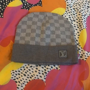 Grå rutig mössa från Louis Vuitton - Snygg grå mössa från Louis Vuitton med ett klassiskt rutigt mönster. Mössan har en uppvikt kant med en broderad LV-logga. Perfekt för att hålla sig varm med stil.