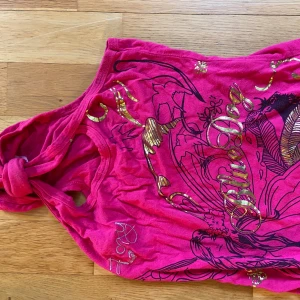 Rosa ärmlös topp med guldtryck - Säljer en rosa ärmlös topp med ett iögonfallande guldtryck och svart mönster. Toppen har en knutdetalj på ena axeln och texten 'Blue' i guld. Perfekt för en färgglad och unik stil.