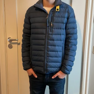 Blå pufferjacka från Parajumpers - Säljer en snygg blå pufferjacka från Parajumpers med dragkedja och en cool patch på ärmen. Jackan har en normal passform och är perfekt för kyligare dagar. Den quiltade designen ger både stil och värme. Storlek 14Y men passar mellan 12-15. Små tecken på användning finns vid ränslar och krage. Går att tvätta såklart.