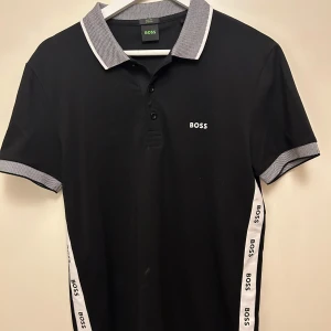 Svart pikétröja från Boss - Snygg svart pikétröja från Boss med vita detaljer och logotyp på bröstet. Knappt använd 