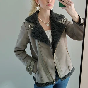 Grå jacka med fuskpälsfoder - Snygg grå jacka med mjukt fuskpälsfoder och dragkedja. Jackan har långa ärmar och bältesdetaljer vid ärmslut och axlar. Perfekt för kyligare dagar med en stilren look. Saknar skärpet som ska sitta längs nedre kanten, var så när den köptes redan men påverkar inte funktionen. 