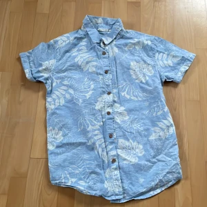 Blå blommig skjorta från KappAhl - Säljer en snygg blå skjorta från KappAhl med ett blommigt mönster i vitt. Skjortan har korta ärmar och knappar framtill. Perfekt för en avslappnad sommarlook! 🌸