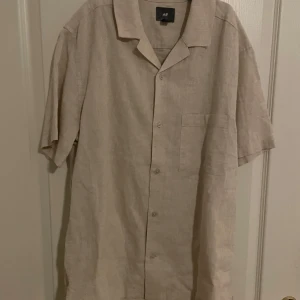 Beige linneskjorta från H&M - Säljer en stilren beige skjorta från H&M i linne. Skjortan har korta ärmar och en klassisk krage med knappar framtill. Perfekt för en avslappnad look.