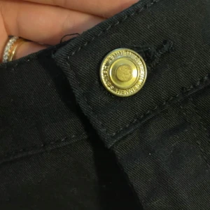 Svarta byxor med knappdetalj - Oanvända tighta svarta byxor från H&M med en knappdetalj i guld. Perfekta för en elegant look. Byxorna har en rak passform och är tillverkade i ett bekvämt material.