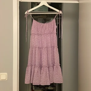 Ljuslila blommig klänning - Charmig ljuslila klänning med blommigt mönster. Klänningen har smala axelband som knyts och en luftig passform. Perfekt för varma dagar och ger en romantisk känsla.