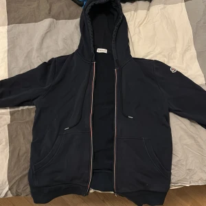 Mörkblå hoodie från Moncler - Säljer en stilren blå hoodie från Moncler med dragkedja och huva. Jackan har långa ärmar och en broderad logga på ärmen. 