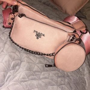 Rosa axelväska från Prada - Snygg rosa axelväska från Prada med justerbar axelrem och en rund avtagbar ficka i mocka material. Man kan även sära väskorna från varann.  Väskan har en kedjedetalj och flera dragkedjor för säker förvaring. Har ej kvitto eller box. Fick i present och är ej säker på att den är äkta. Därav det billiga priset! Och priset kan även diskuteras :) 