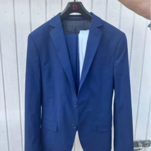 Blå kavaj från Dressmann - Snygg blå kavaj från Dressmann i slim fit. Perfekt för formella tillfällen med två knappar framtill och klassisk design. Passar utmärkt för både vår och sommar.