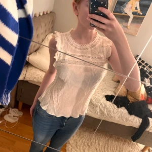 Vit ärmlös blus med spetsdetaljer - Säljer en söt vit ärmlös blus med fina spetsdetaljer och volanger vid ärmhålen. Blusen är använd vilket syns på nära håll då knappen baktill är trasig samt att en liten fläck finns på vänstra armen. Inget som syns på håll.