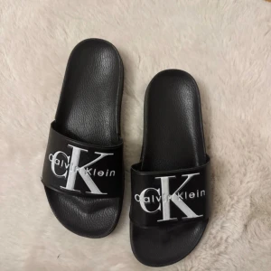 Svarta sandaler från Calvin Klein - Snygga svarta sandaler från Calvin Klein med en bred rem prydd med märkets logotyp i vitt. Perfekta för en avslappnad stil och bekväma att bära.