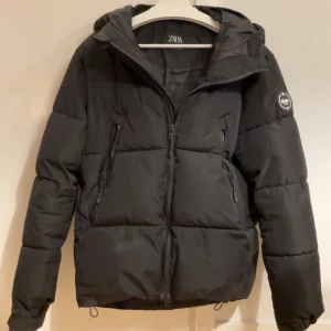 Svart pufferjacka från Zara - Snygg svart pufferjacka från Zara med dragkedja och huva. Jackan har en quiltad design och långa ärmar. Perfekt för kyliga dagar och för en trevlig skidresa! Hör av er vid frågor. 