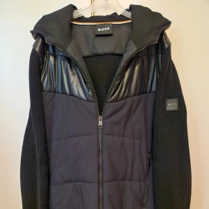 Sommar25. Cardigan jacka från BOSS storlek M - Snygg svart jacka från BOSS med dragkedja och huva. Nypris: 2899:-. Jackan har en quiltad design på framsidan och baksidan, samt stickade ärmar för extra komfort. Två fickor framtill med dragkedja. Använd sparsamt fåtal gånger. Perfekt för kyligare dagar. 