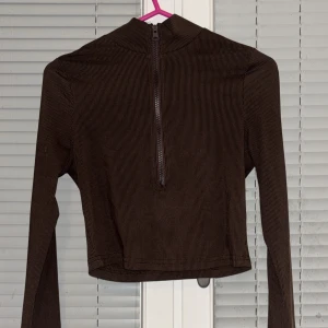 Brun ribbad tröja med dragkedja - Säljer en brun ribbad tröja med dragkedja framtill. Tröjan är croppad och har långa ärmar, perfekt för en stilren look. Passar bra till både jeans och kjol.