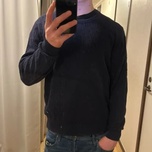 Mörkblå stickad tröja från J.Lindeberg - Säljer en stilren mörkblå stickad tröja från J.Lindeberg. Tröjan har ett diskret kabelstickat mönster och ribbade muddar vid ärmslut och nederkant. Perfekt för en avslappnad men ändå elegant look. 🧥