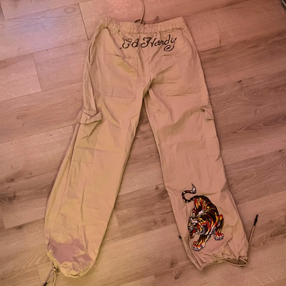 Snygga beige byxor från Ed Hardy med coola tryck och dragsko i midjan. Byxorna har en avslappnad passform och är dekorerade med en tiger på ena benet samt textdetaljer. Perfekta för en avslappnad stil.. Farkut & Housut.