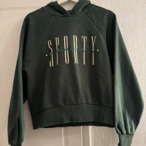Grön hoodie från Gina Tricot - Säljer en grön hoodie från Gina Tricot med texten 'SPORTY' tryckt på framsidan. Tröjan har långa ärmar och en bekväm huva. Perfekt för en avslappnad stil.