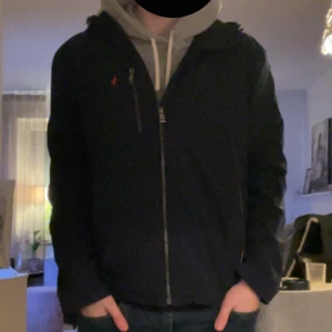 Mörkblå jacka från Polo Ralph Lauren - Säljer en snygg mörkblå jacka från Polo Ralph Lauren med dragkedja och broderad röd logga på bröstet. Jackan har en stilren design med långärmade ärmar och en praktisk bröstficka med dragkedja. Perfekt för en casual look.