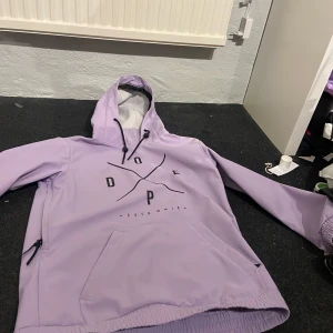 Lila hoodie från Dope - Säljer en snygg lila hoodie från Dope med svart tryck på framsidan. Den har en stor ficka framtill och justerbar huva med svarta snören. Perfekt för en avslappnad stil.