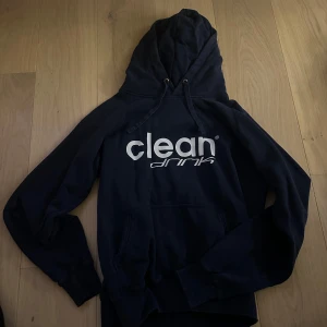 Marinblå hoodie från Clean Drink - Säljer en svart hoodie från Clean Drink med vit logga på bröstet. Den har en klassisk design med känguruficka och justerbar huva. Perfekt för en avslappnad stil.