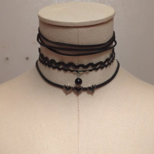 Svart chokerhalsband med hjärtan - Säljer ett set med tre svarta chokerhalsband. Det ena är ett enkelt band, det andra har ett vågigt mönster med en svart pärla, och det tredje har små silverfärgade hjärtan. Perfekt för att ge en edgy touch till din outfit!